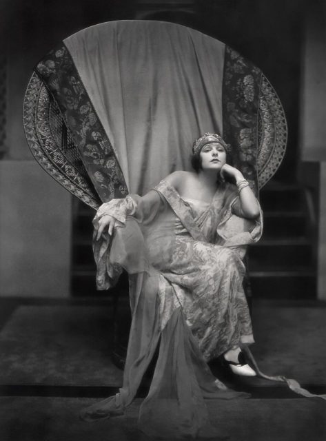 Dramatic publicity shot of actress Norma Talmadge seated in a large wicker peacock chair.