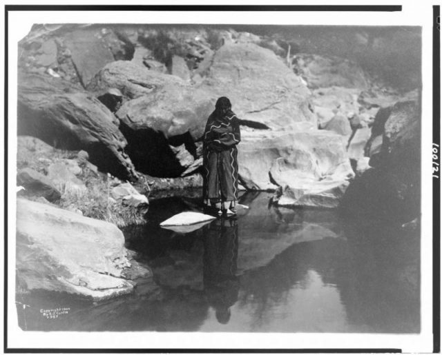Navajo Woman standing on rock, reflected in a pool of water. Her skirts would have been woven from yucca fiber.