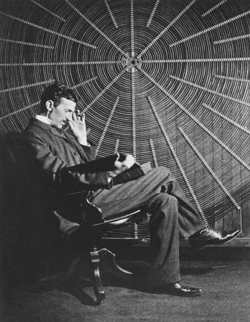 Tesla sitting in front of a spiral coil used in the wireless power experiments at his East Houston St. laboratory.