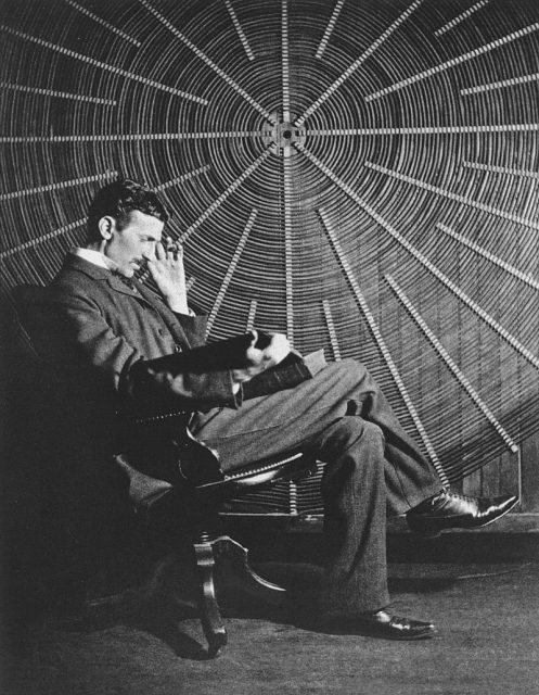 Tesla sitting in front of a spiral coil used in his wireless power experiments at his East Houston St. laboratory.