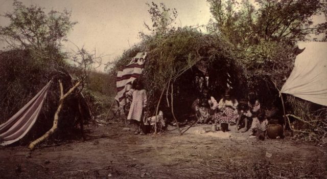 A Chiricahua Apache medicine man sits with his family at their brush-made wickiup. This is also at a camp in Arizona.