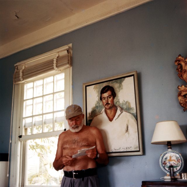 Ernest Hemingway at his home in Cuba, c. 1953, standing in front of a 1929 portrait of himself by Waldo Pierce.