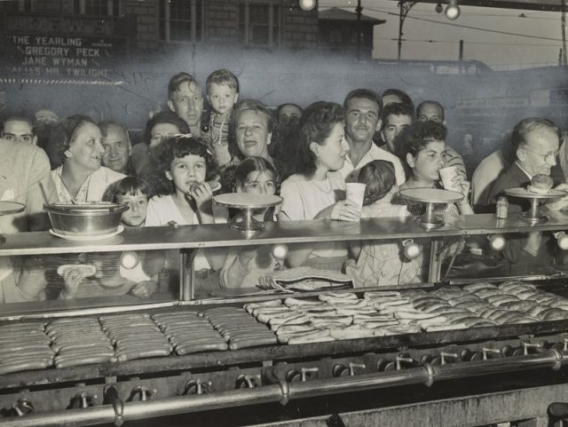 Photograph showing a hungry crowd eating and drinking at Nathan’s food counter with hot dogs and rolls cooking on the grill.