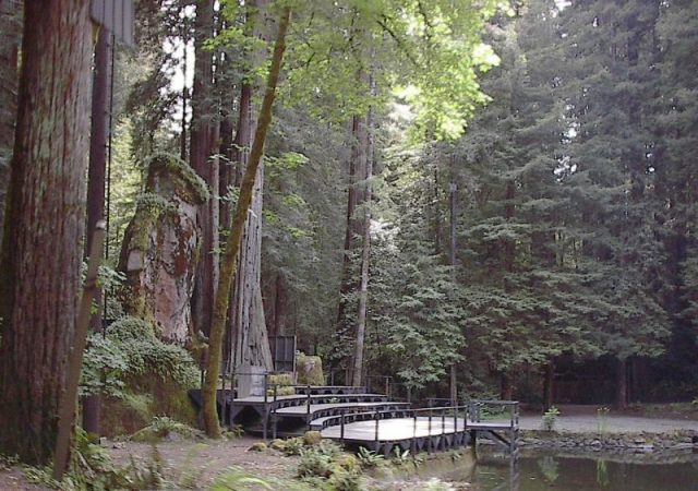 The Owl Shrine covered in moss, standing among trees behind a stage at one edge of a man-made pond. Photo by Aarkwilde CC BY-SA 3.0