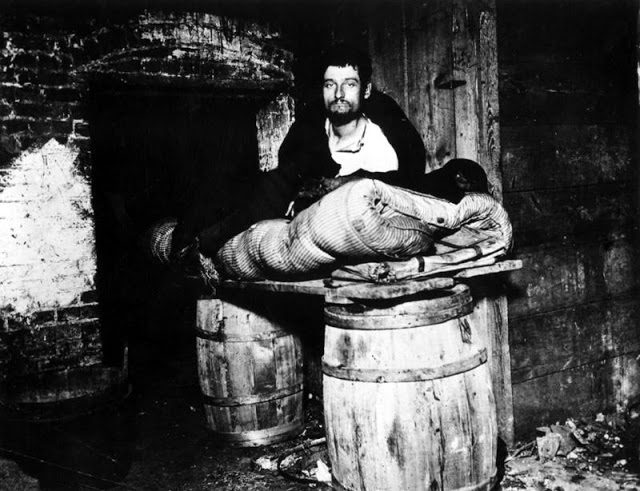 A peddler sits on his bedroll, atop two barrels, in the filthy cellar he lives in. New York, 1886.