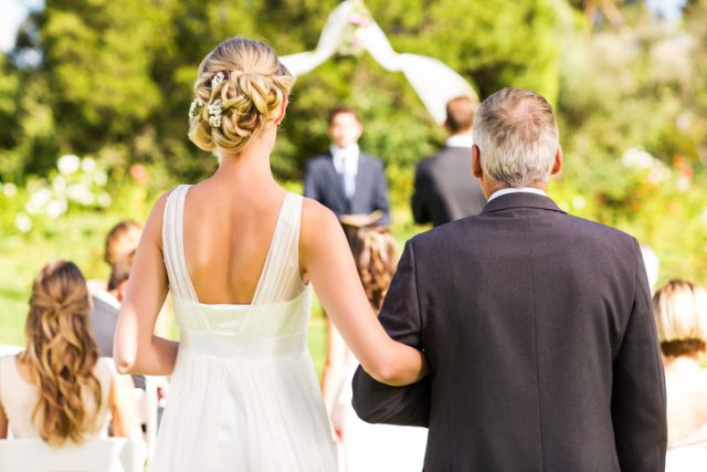 Rear view of bride and father walking down the aisle with people in background during outdoor wedding. Horizontal shot.