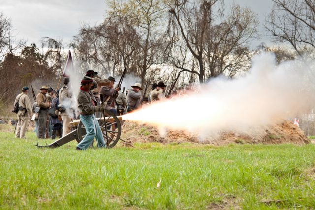 Men dressed in period costume of the 1800s portray soldiers from the Civil War during a reenactment of the Battle of Big Bethel in Newport News, Virginia, which took place in 1861. Here the cannon is caught mid-blast with the surrounding soldiers covering their ears to protect their hearing from the loud explosion.
