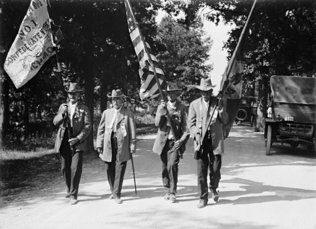 Four men walking down a street, with three of them carrying flags on flagpoles