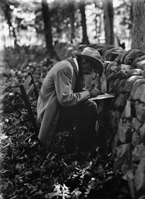 Man writing on a piece of paper while seated next to a stone fence