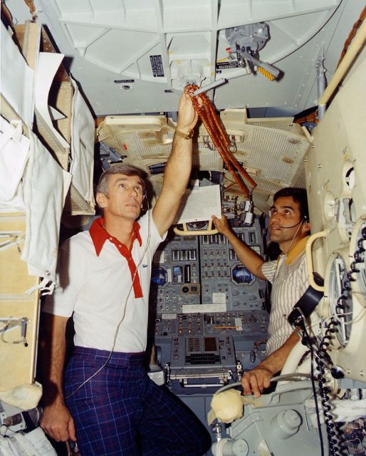 Apollo 17: Inside the Lunar Module training simulator, Gene Cernan drapes the cords for the LM cabin utility lights on the handle of the rendezvous hatch. The two utility lights are just to the right of Jack Schmitt’s right hand and are attached to the Alignment Optical Telescope (AOT) guard with clamps.