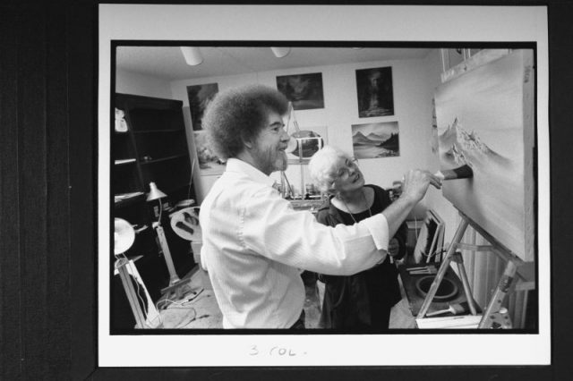 TV painting instructor/artist Bob Ross at easel painting one of his mountain landscapes as his business partner Annette Kowalski looks on.
