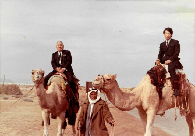 Man standing between Chiune and Nobuki Sugihara, who are sitting on the backs of camels