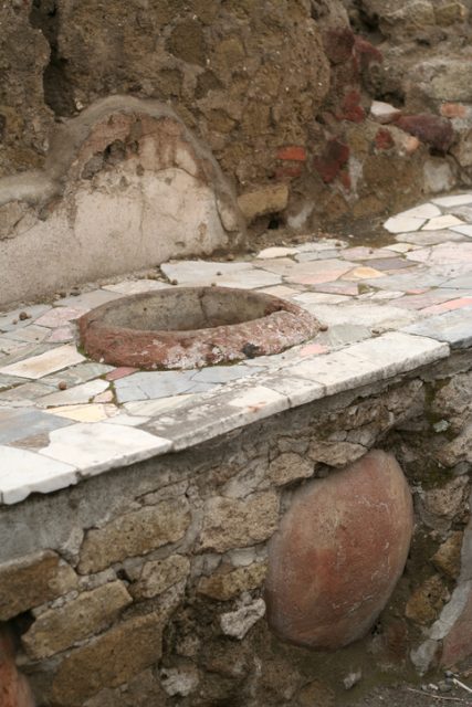 “A thermopolium was a place, used during the times of ancient Rome were it was possible to purchase ready-to-eat food. It was made of a small room with a front desk, and hot food was stored in big clay pots inserted in the front desk. It was probably used in a way similar to modern fast foods. Remains of thermopolia are preserved in Pompeii and Herculeanum.”