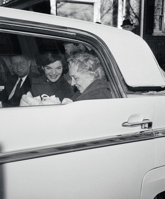 Jackie and John F. Kennedy riding in the back of a car with Maud Shaw, who's holding a newborn John F. Kennedy, Jr.
