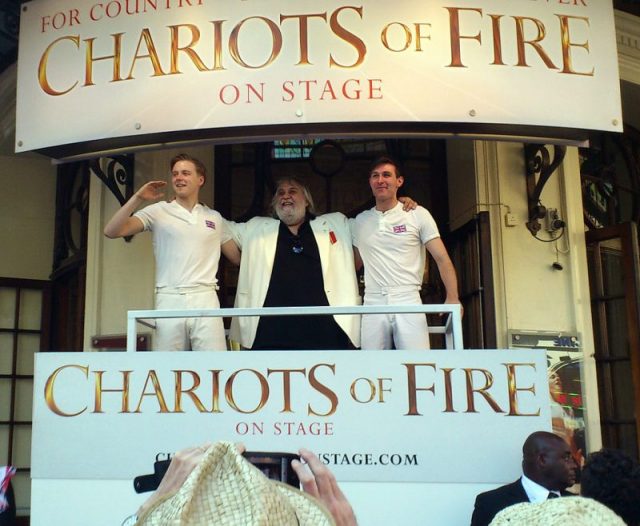 The Chariots of Fire stage adaptation: Stars Jack Lowden and James McArdle flank Vangelis, watching the Olympic Torch Relay set to the iconic tune, from the Gielgud Theatre, July 2012. Photo by Markdawson7 – CC BY-SA 3.0