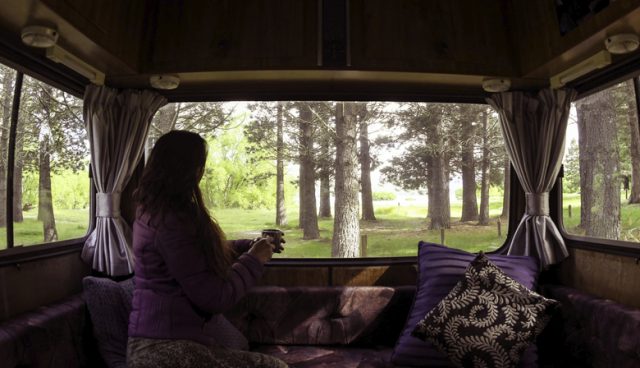 A woman sits in her motor home lounge, warm cup of coffee in her hands, looking out the rear window at the beautiful forest that surround her, lit by the morning sunlight.