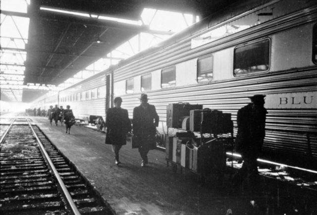 A man and a woman walking with a porter on the platform next to a railroad passenger car at a train station