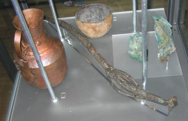 Finds from a vǫlva’s grave in Köpingsvik, Öland. There is an 82 cm long wand of iron with bronze details and a unique model of a house on the top. There is also a pitcher from Persia or Central Asia, and a West European bronze bowl. Dressed in a bear pelt, she had received a ship burial with both human and animal sacrifice. The finds are on display in the Swedish History Museum in Stockholm. Photo by Berig CC BY-SA 4.0