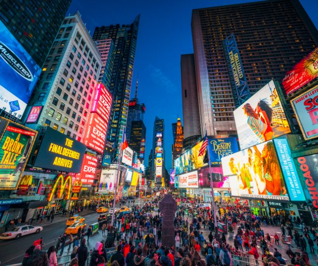 Times Square covered with illuminated billboards during the ‘blue hour’ period at dusk, with tourists on the streets.