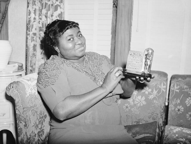 Hattie McDaniel holding up her Oscar while sitting on a couch