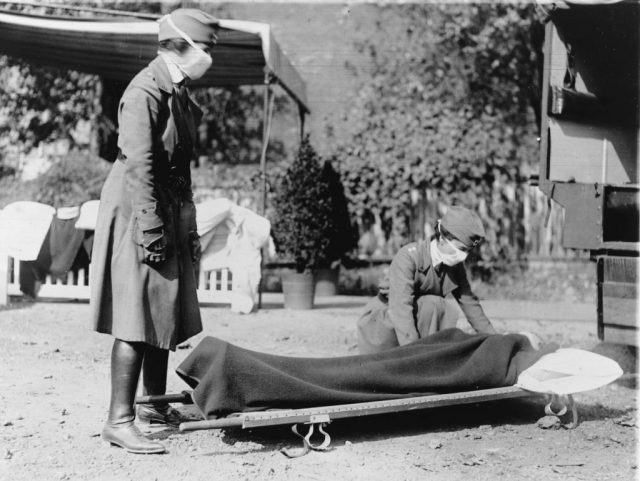 Two Red Cross nurses standing over a stretcher on the ground outside
