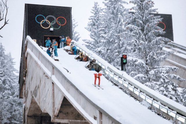 Olympic skier going down a snow-covered ramp while other athletes watch