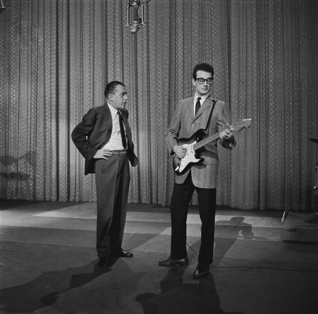 Ed Sullivan standing next to Buddy Holly while he plays guitar