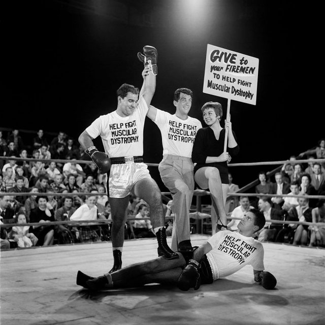 Dean Martin standing over Jerry Lewis in a boxing ring while a woman holds up a sign in support of Muscular Dystrophy awareness