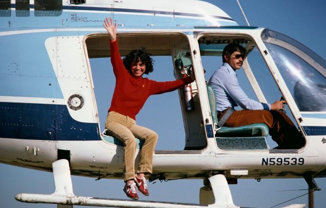 A stunt performer, playing Lynda Carter in Wonder Woman, waves from a helicopter before jumping.