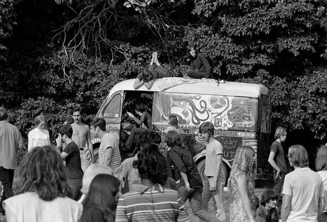 A group of hippies gathered around a decorated van in the woods