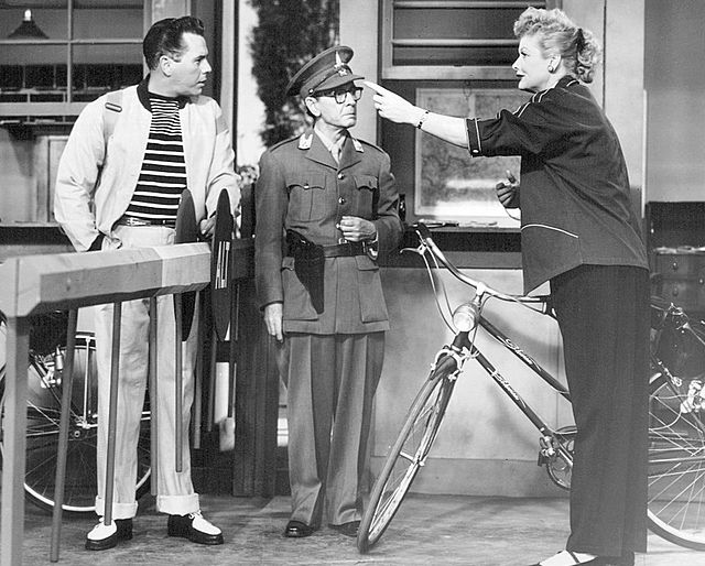 Lucille pointing at the hat of a man in uniform while Desi watches from behind a low fence