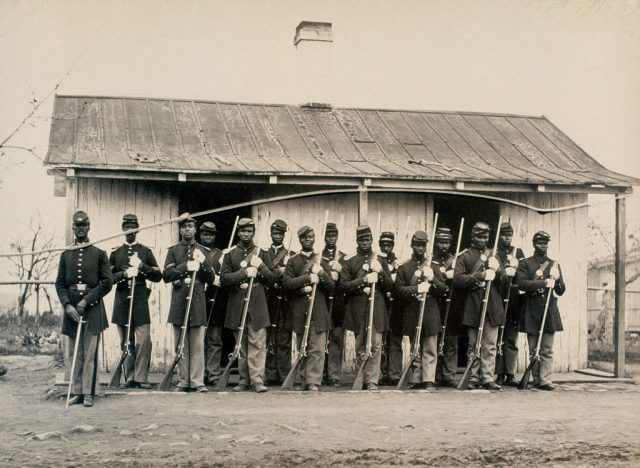 15 soldiers with the 107th United States Colored Troops standing in a row with rifles in their hands