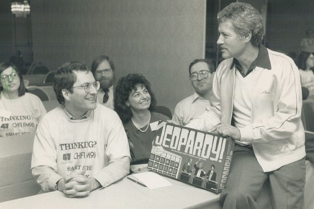 Alex Trebek sitting on a table while people look at him