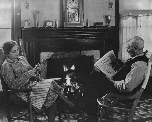 A farmer reads a newspaper in front of the fire as his wife cuts up some material in South Carolina circa 1940s. (Photo Credit: FPG/Getty Images)