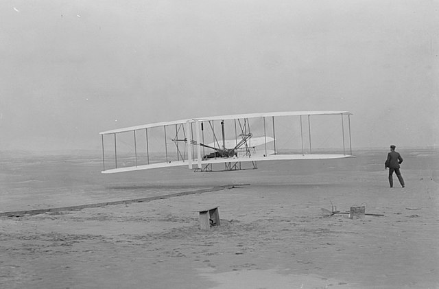 Orville Wright sitting in an airplane while Wilbur Wright looks on