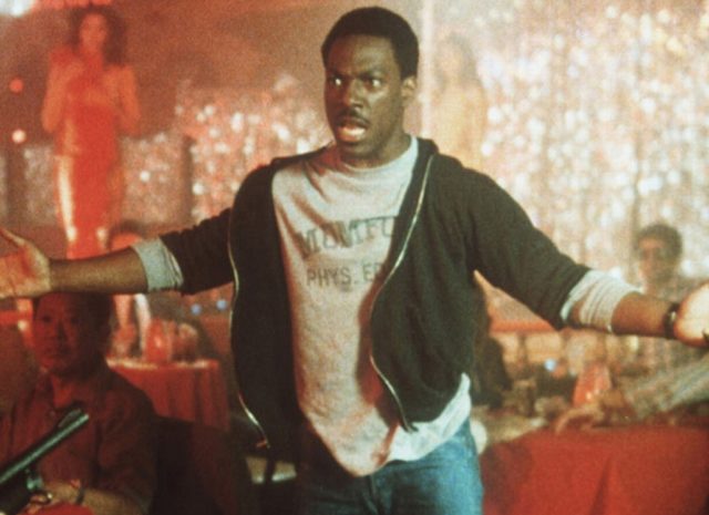 Axel Foley standing with a shotgun pointed at him