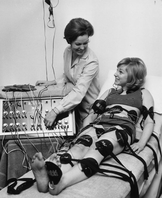 Paula Byrne demonstrates an electronic slimming device to actress Alison Frazer. Pads all over Alison’s body stimulate her muscles with a mild electric current, with the same result as exercise. (Photo Credit: Keystone/Getty Images)