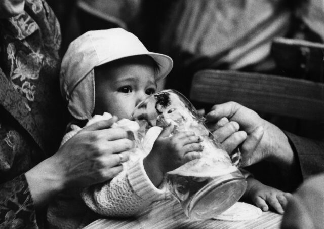 A baby drinking beer from a glass.