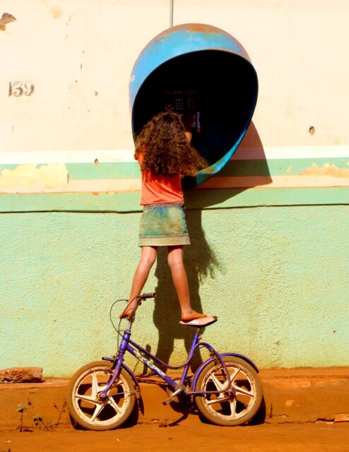 A child balances on a bike as she uses a payphone.