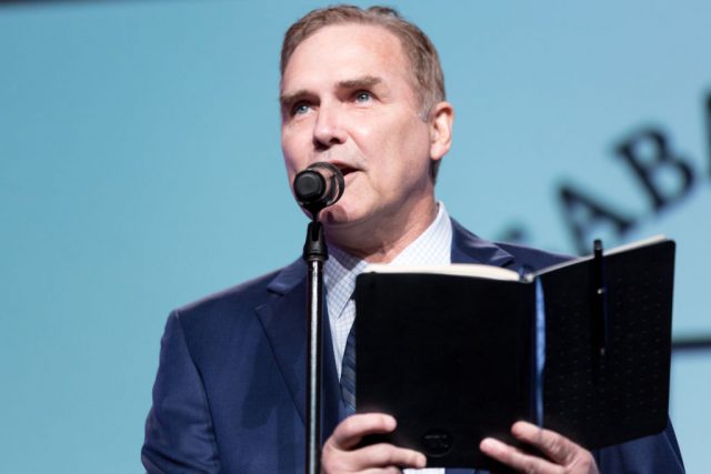 Norm Macdonald holding a book and standing behind a microphone