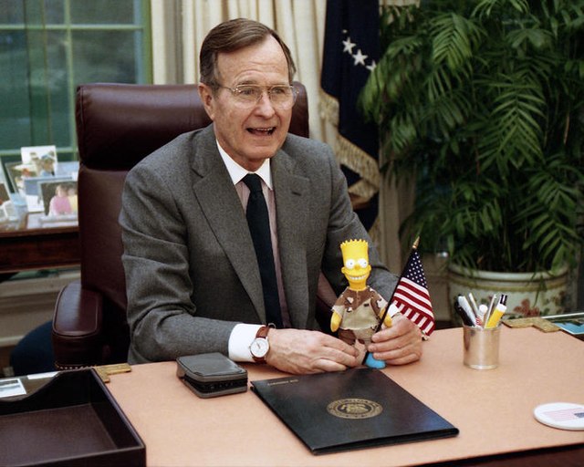 President Bush sits at his desk in the Oval Office and holds a Bart Simpson doll.  (Photo Credit: George Bush Presidential Library and Museum, accessed via Wikimedia Commons)