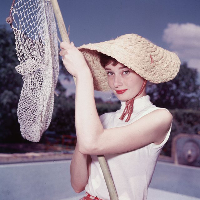 Portrait of Belgian-born American actress Audrey Hepburn (1929 – 1993) as she wears a peculiar hat and sleeveless blouse and holds a pool cleaning net beside a dry swimming pool, early 1950s. (Photo Credit: Hulton Archive/Getty Images)