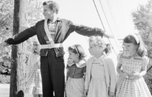 Male School Safety Patrol member standing before a group of children with his arms outstretched