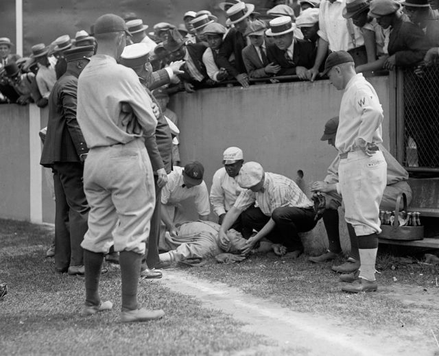Pro baseball star Babe Ruth lies unconscious on the ground surrounded by medics, players, and fans.