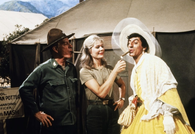 Harry Morgan, Loretta Swit, and Jamie Farr in character while filming "M*A*S*H", Swit lights a cigar for Farr who is dressed as a woman.