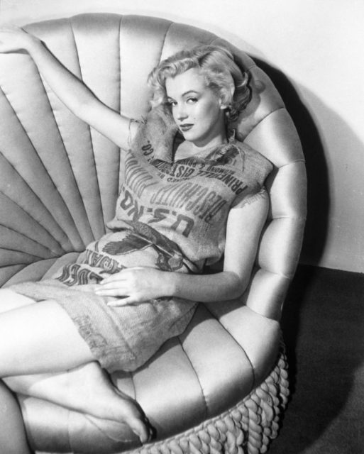 Marilyn Monroe stares into the camera while posing on a chair in the potato sack dress.