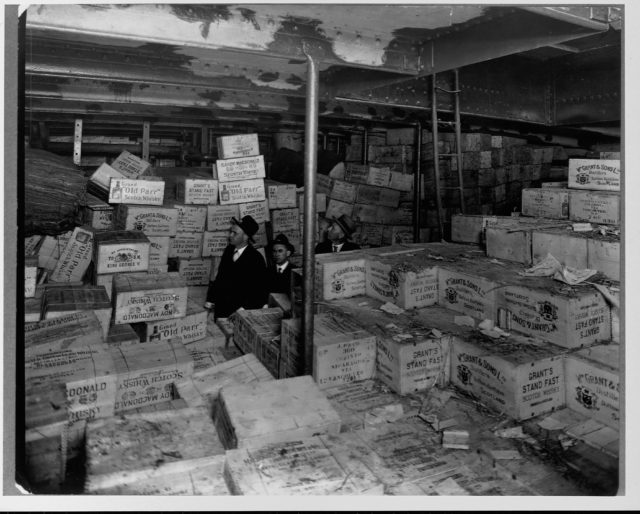 Two men standing among crates of confiscated alcohol during prohibition