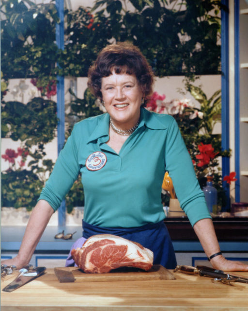 Portrait of Julia Child in turquoise behind a slab of meat in her kitchen