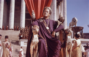 Coloured photo of Rex Harrison playing Julius Caesear, sitting in a throne wearing purple robes.