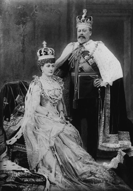 Black and white photo of a man standing beside a sitting woman. Both of them wear extravagant, regal clothing and crowns.